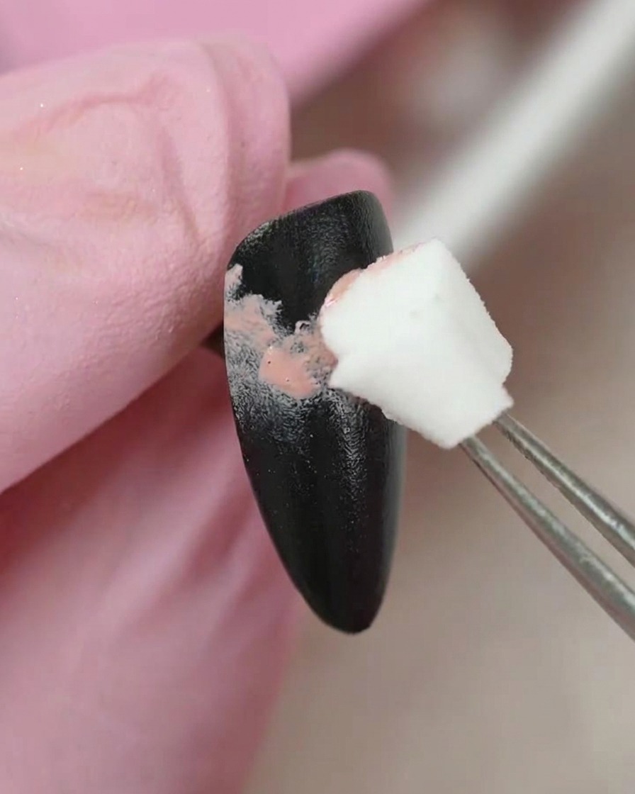 A pair of tweezers holding a small sponge applicator, gently dabbing pink gel color onto a black-painted nail surface.