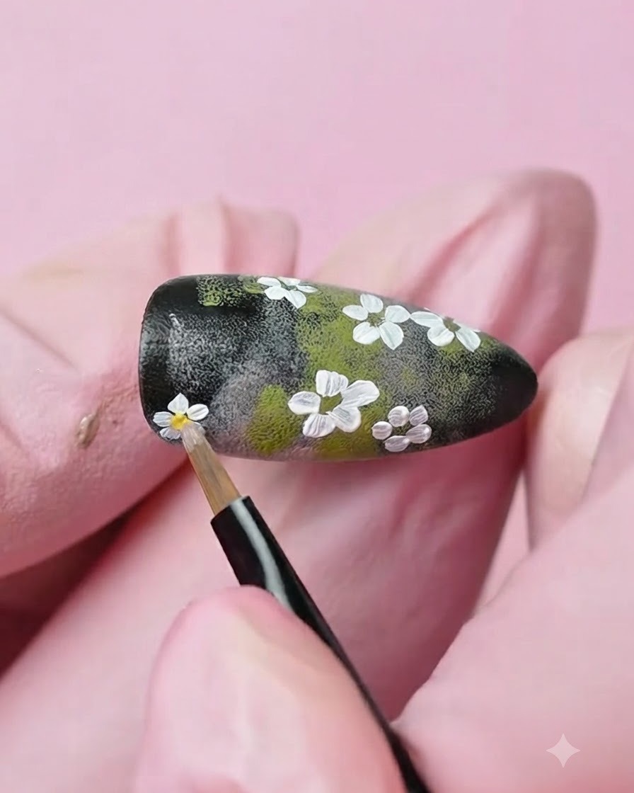 A fine detail brush applying a dot of yellow gel to the center of a painted white flower on the nail.