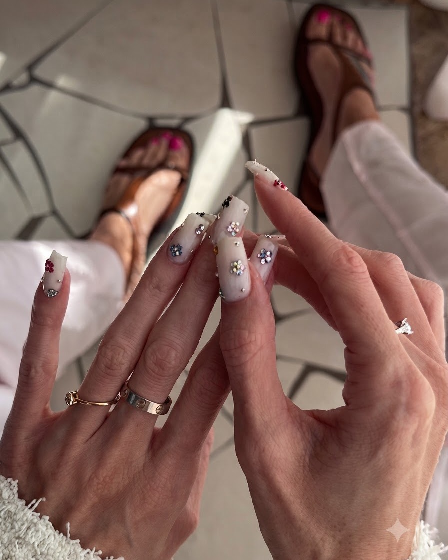 Close-up view of square milky white nails decorated with colorful floral charms on a neutral background.
