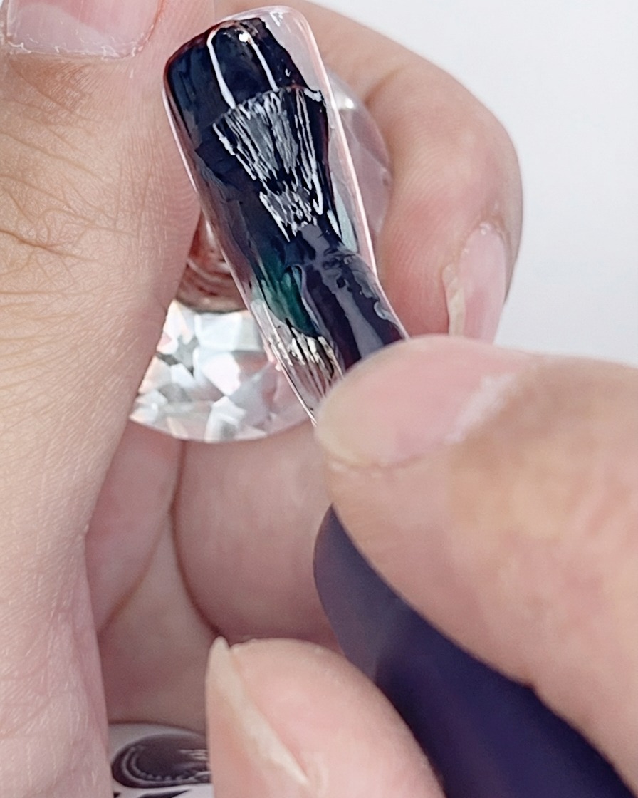 A brush applying the first coat of black gel polish onto an artificial nail tip.