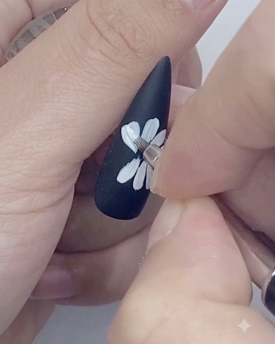 A fine liner brush tracing the outer edges of white daisy petals painted on a black nail.