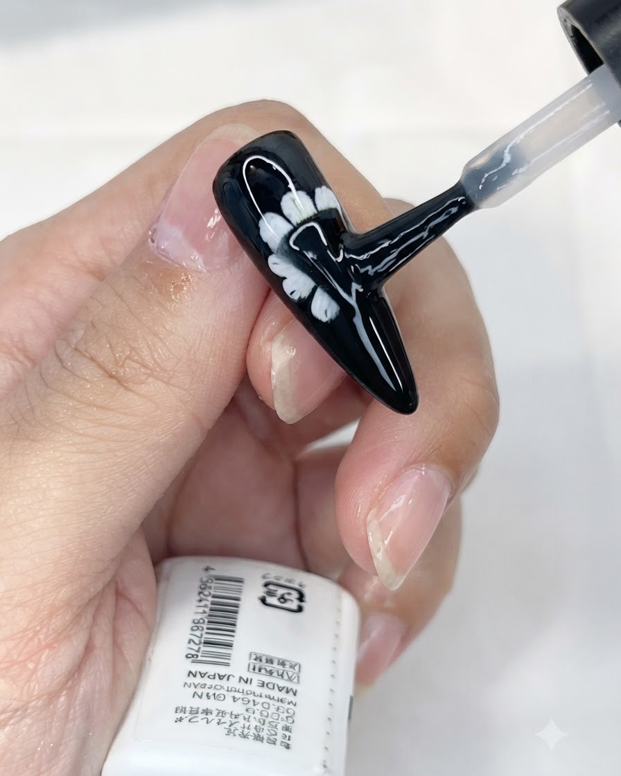 A brush applying a layer of matte top coat over a black nail with white daisy art.