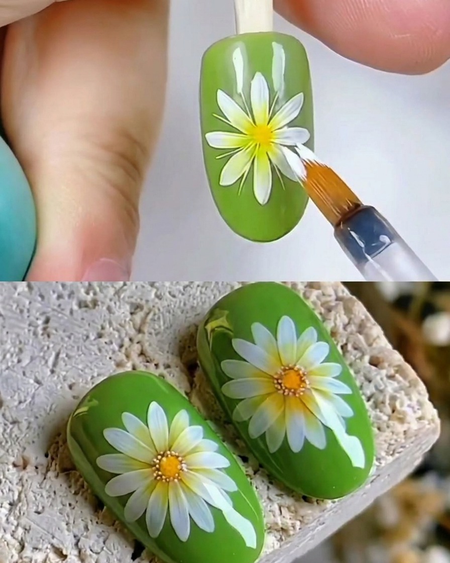 A close-up of a brush applying a glossy clear top coat over a white daisy nail art design on a green nail.