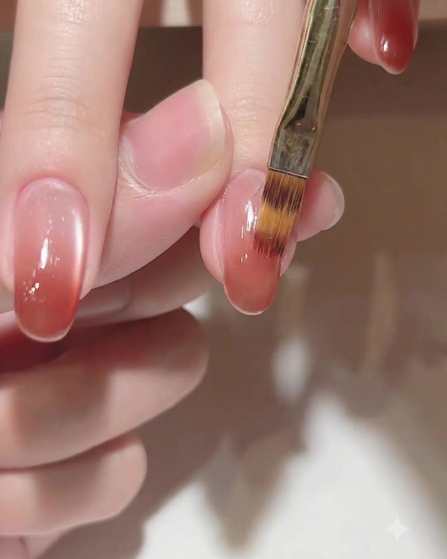 A close-up of a brush gently feathering a warm pinkish-brown gel color gradient toward the cuticle on an oval nail.