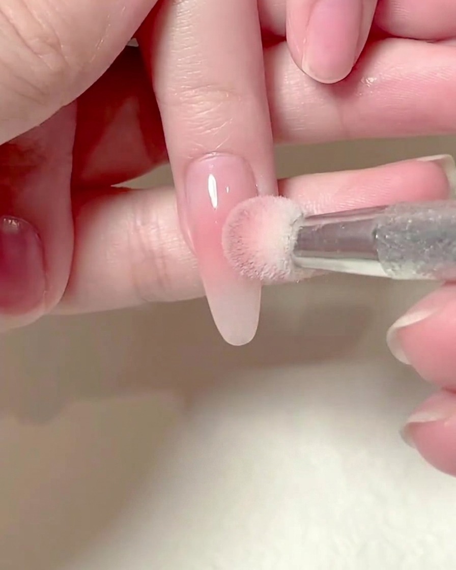 A square sponge held by a hand gently patting pink blush gel onto an elongated fingernail.