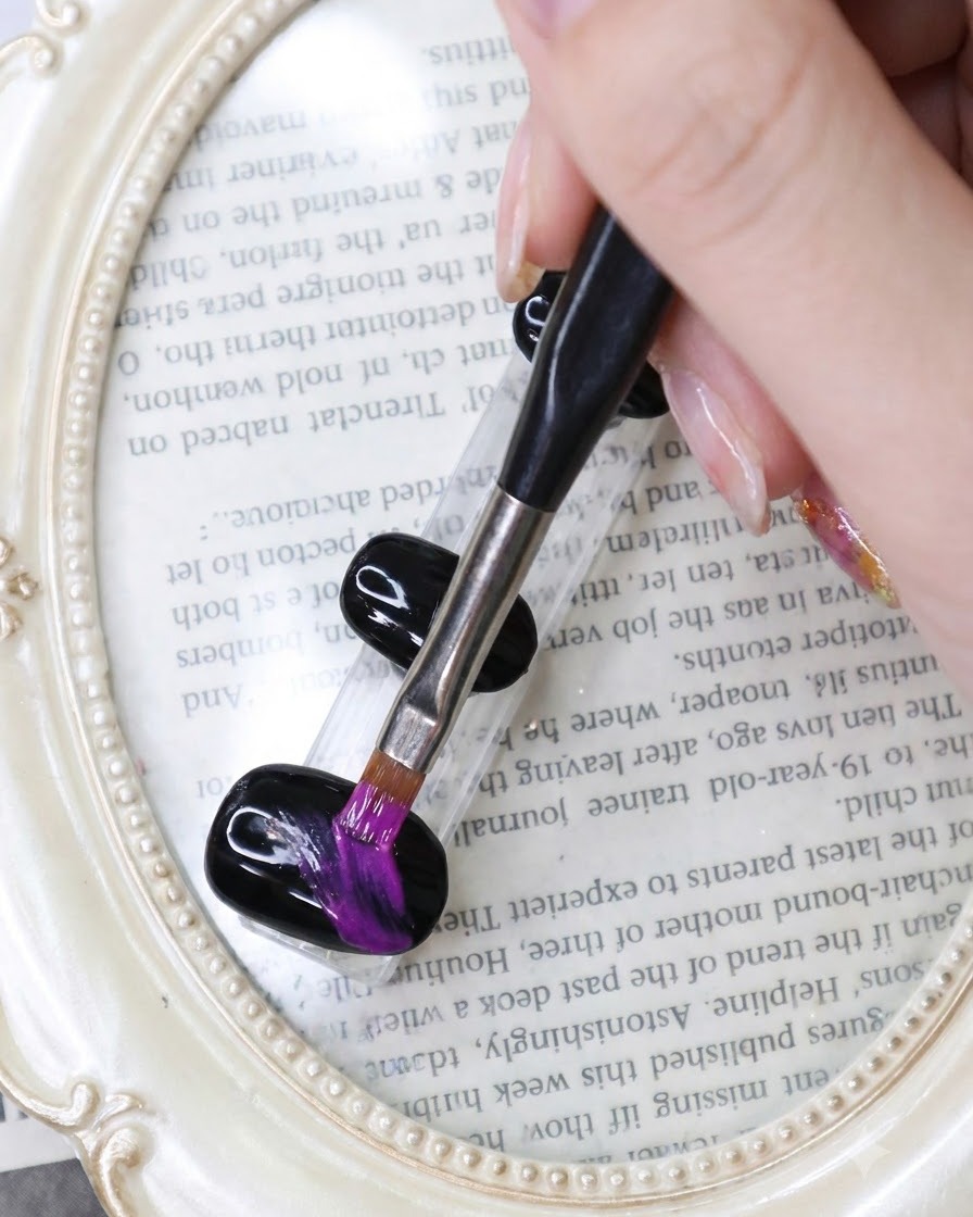 A fine liner brush drawing vibrant neon purple strokes across a black gel nail on a book-themed background.