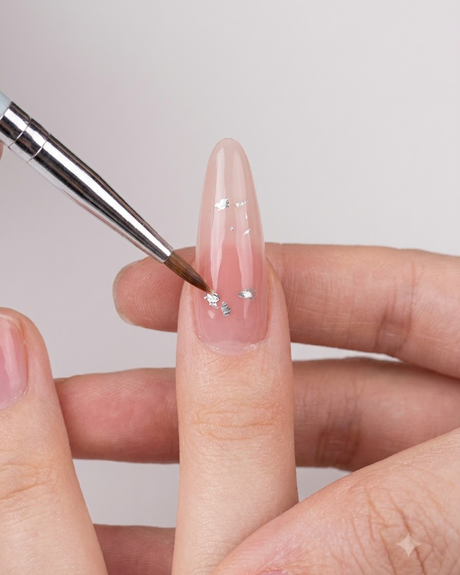 A fine nail art brush placing small silver iridescent flakes onto a nude glossy fingernail.