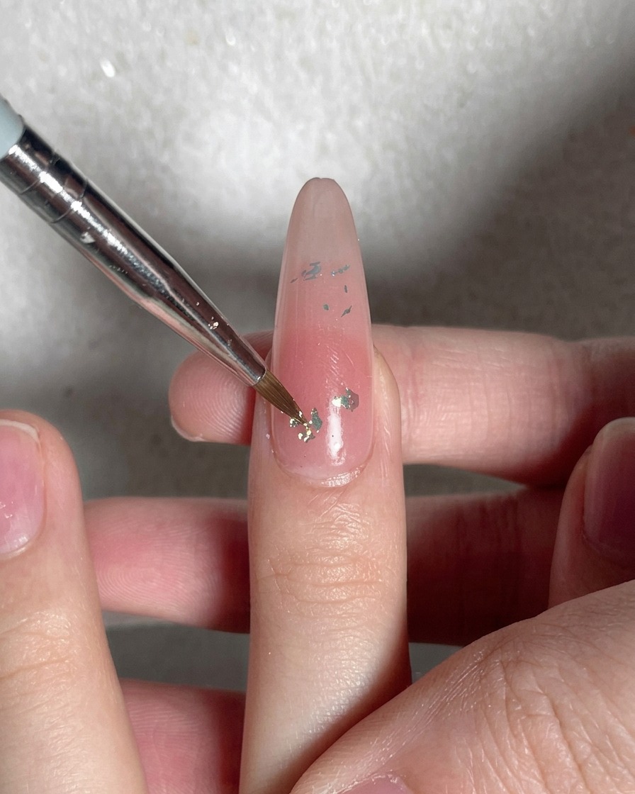 A detail shot of a brush tip pressing down flatly onto iridescent flakes on a nude nail.