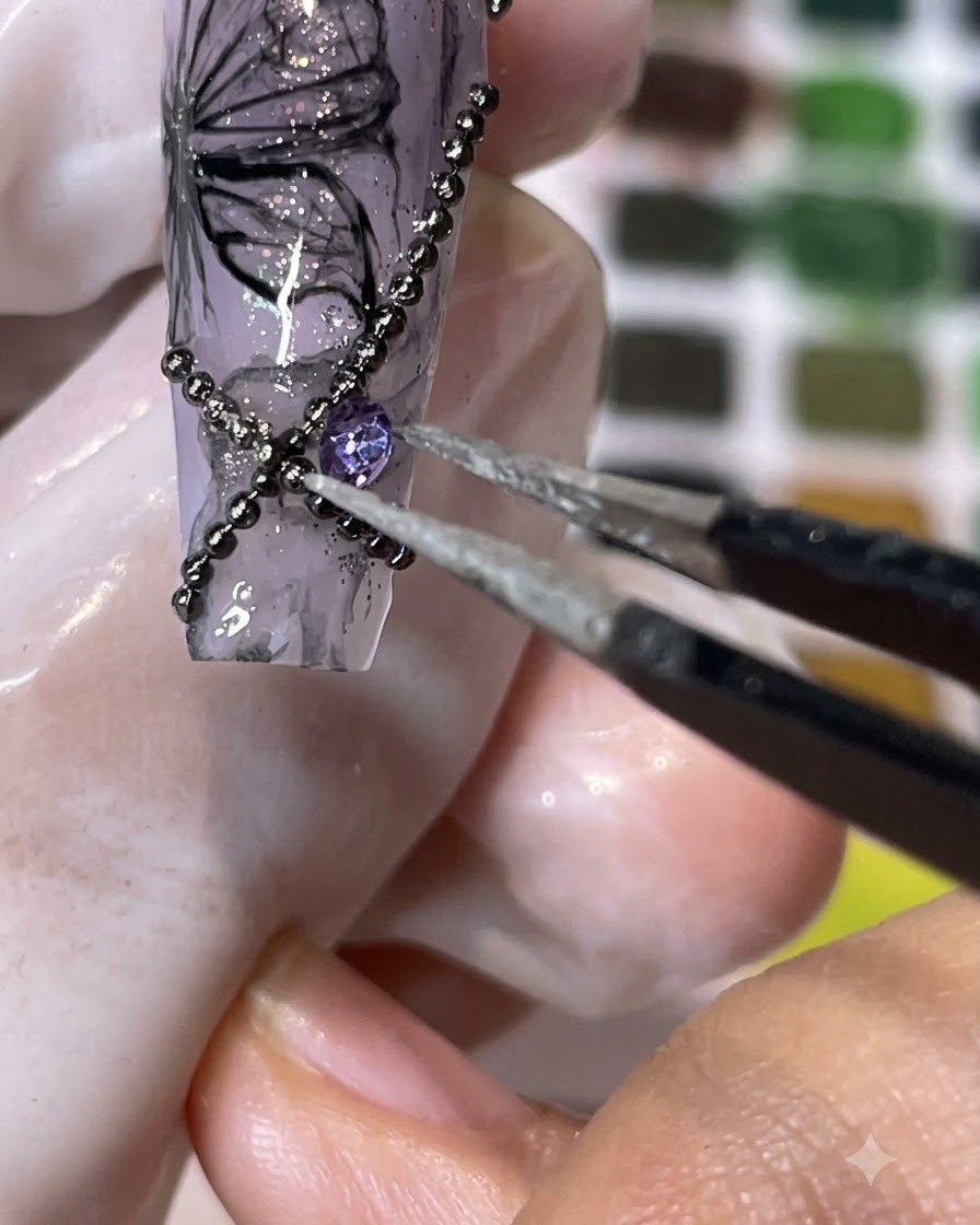A pair of tweezers placing a purple pointed-back rhinestone onto a decorated nail surface.
