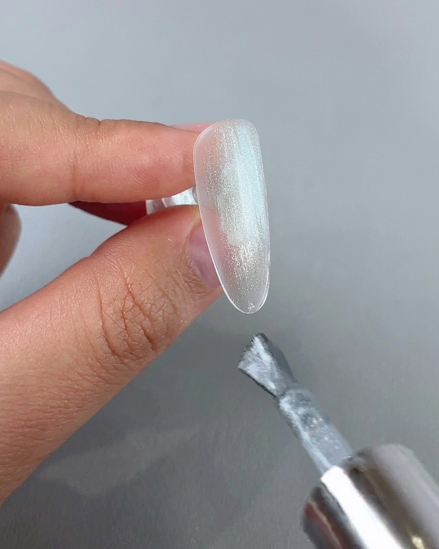 A nail brush applying a coat of sparkly silver cat eye gel onto a clear nail tip.