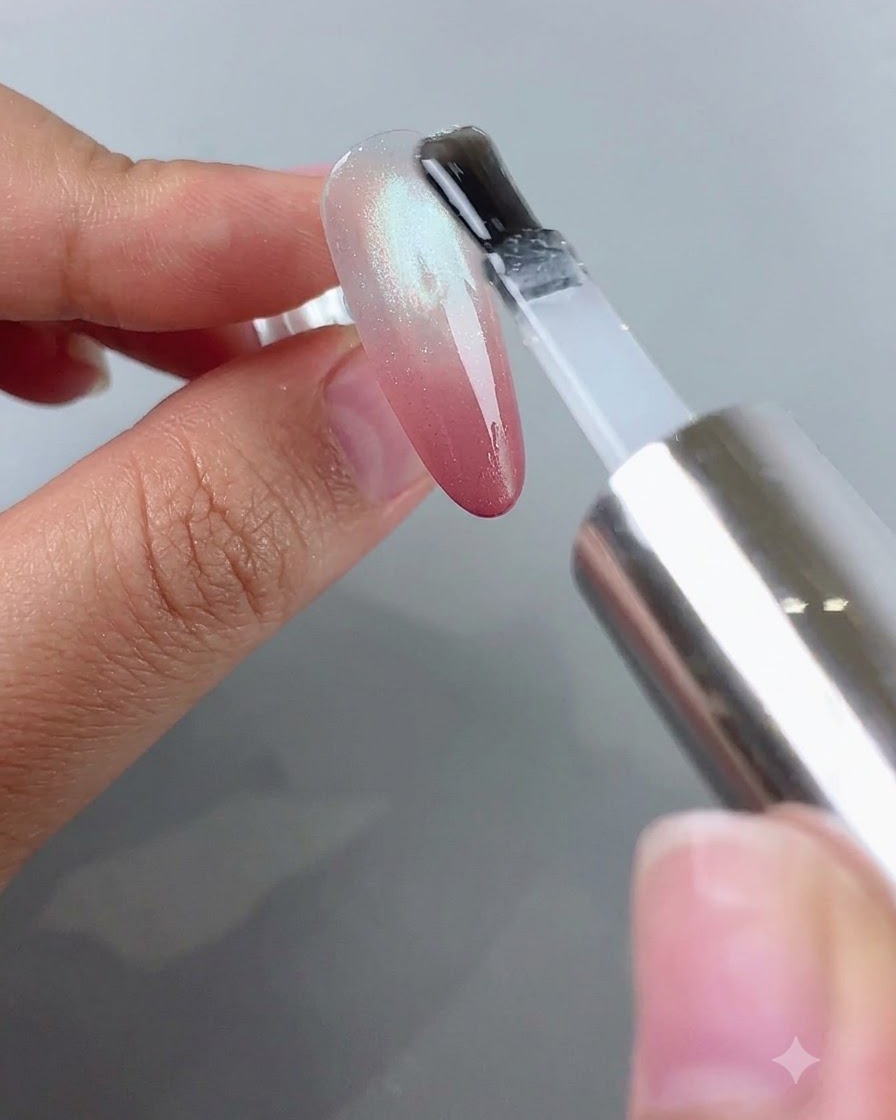 A brush applying clear top coat over a nail that has a pink gradient blush and silver cat eye effect.