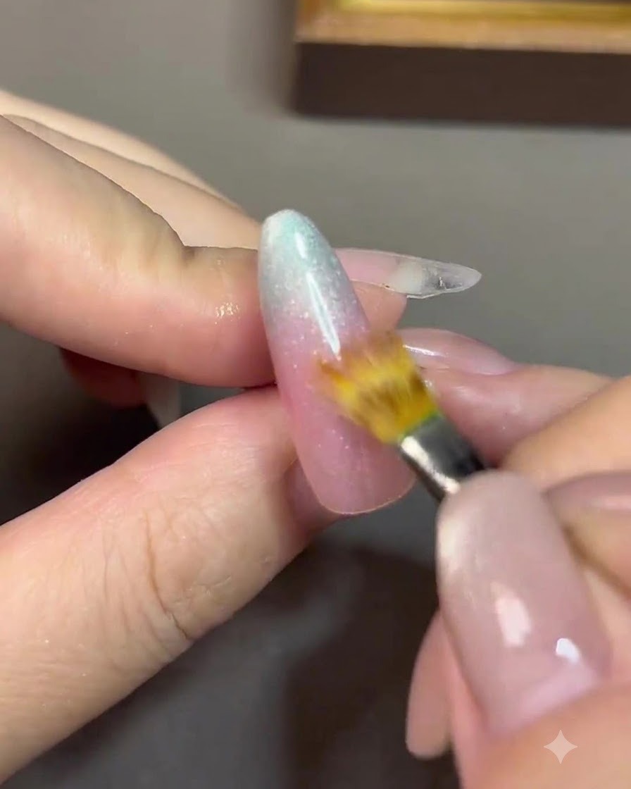 A fine detail brush blending the transition line between a nude pink gel and a blue gel on a nail.