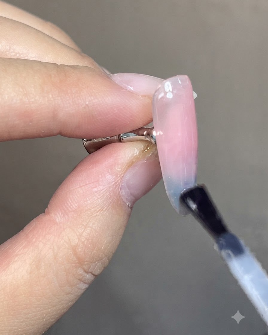 A brush applying a clear glossy top coat over a finished blue and pink cat-eye gradient nail design.