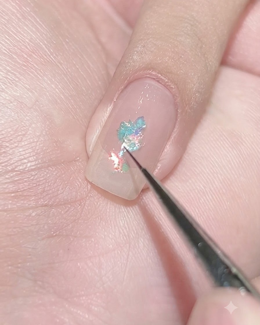 A fine liner brush depositing tiny light blue flakes onto a clear-coated nail to create a concentrated cluster.