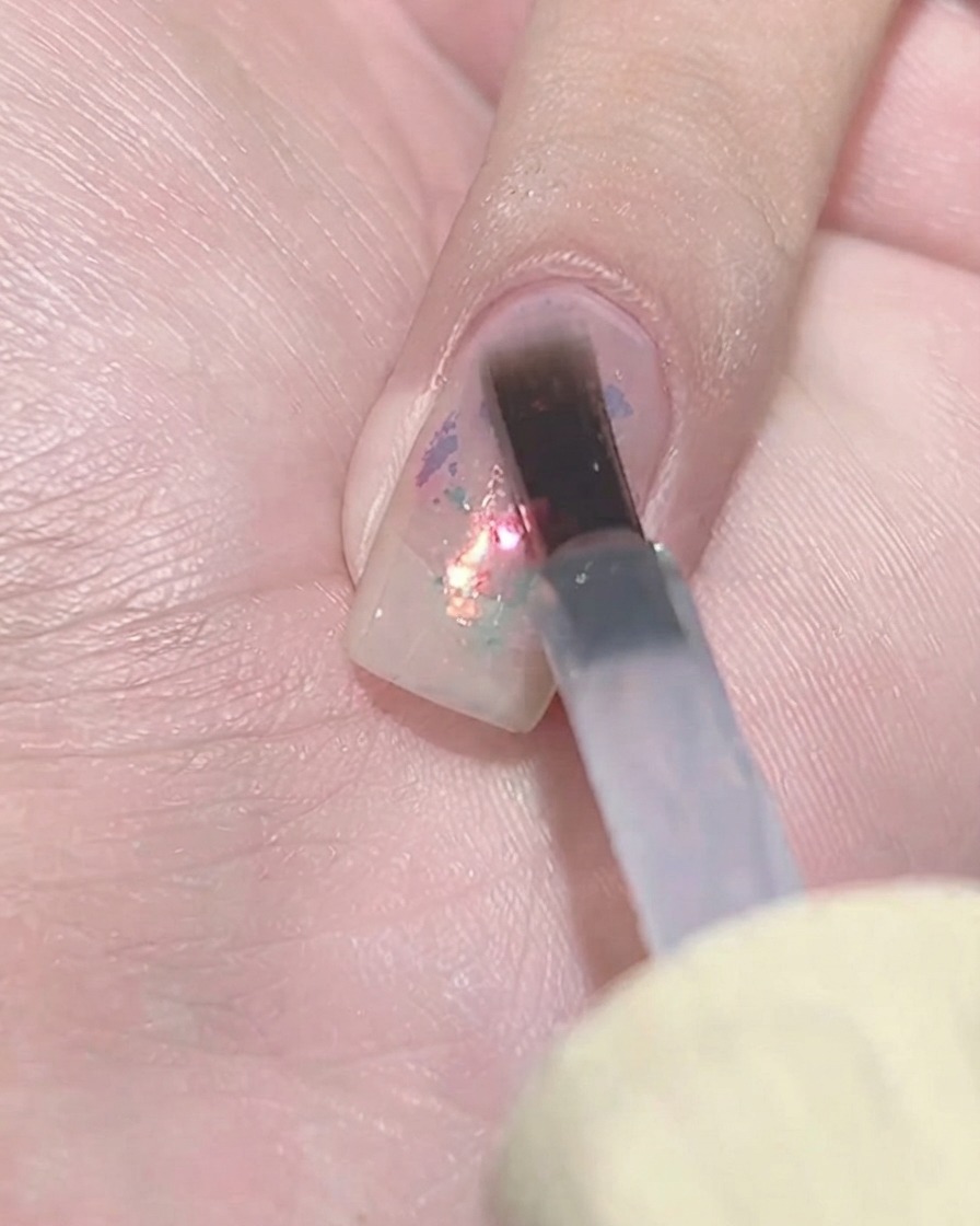 A flat gel brush dabbing a layer of sheer purple polish over a textured flake cluster on a fingernail.