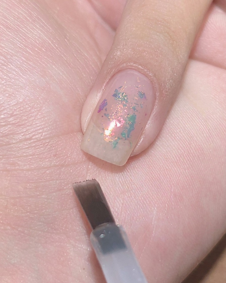 A flat gel brush hovering near a clear nail with embedded flakes, ready to dab sheer purple polish.