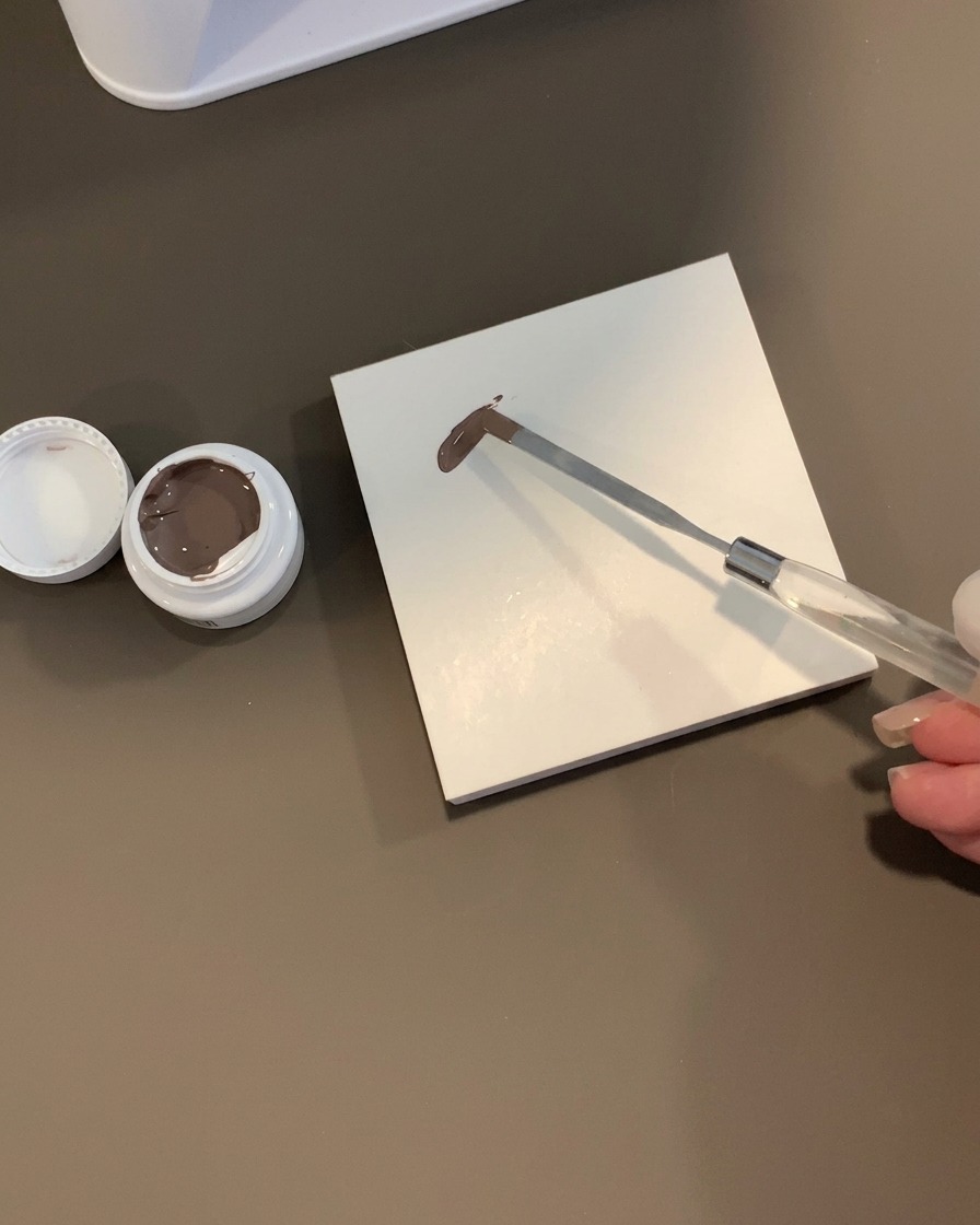 A metal spatula holding a dollop of brown-grey gel on a white square tile palette next to an open jar.