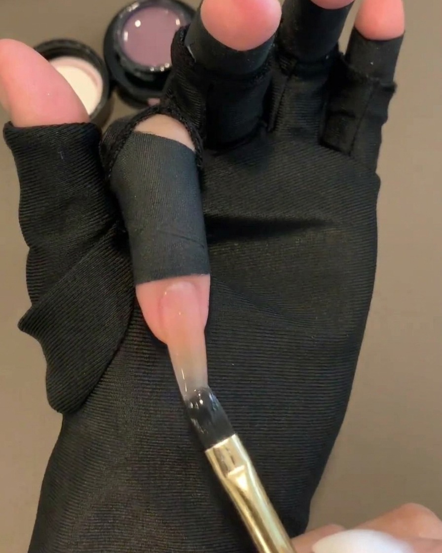 A flat gel brush applying a sheer purple color gel to the upper half of a nude base nail.