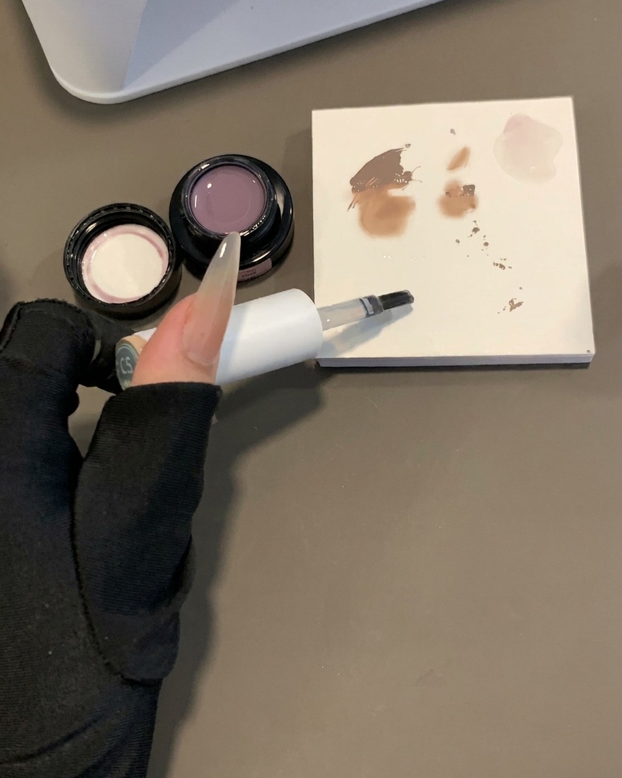A gel brush being dipped into clear base gel next to a mixing palette with pigment swatches.