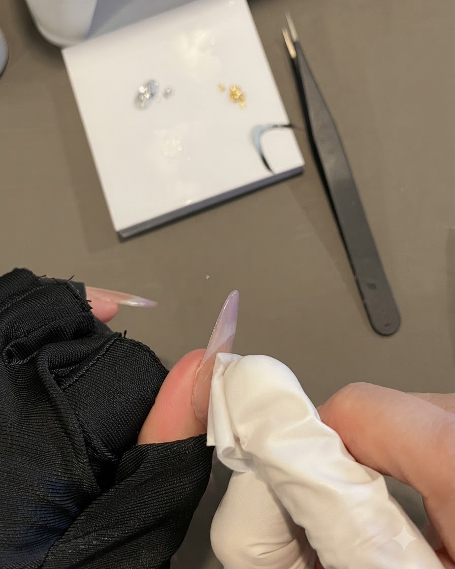 A gloved hand wiping a nail surface with a lint-free wipe while holding a long oval nail.