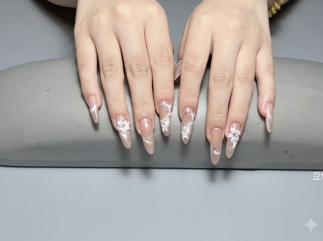 A close-up shot of long almond nails featuring sculpted white 3D flowers and tiny silver crystals on a glossy nude base.