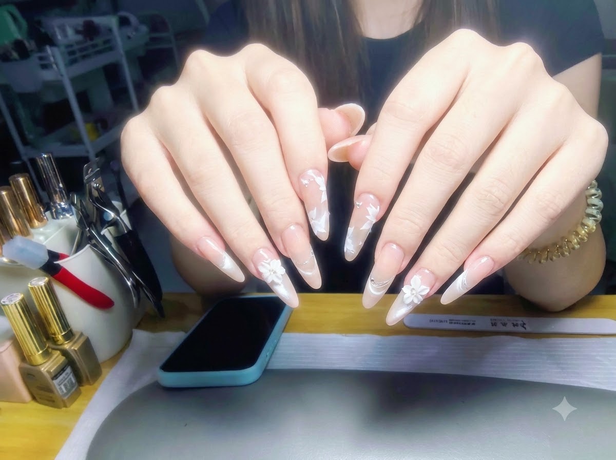 Both hands held up against a workstation background, displaying a full set of glossy nude pearl gradient nails with white floral art.