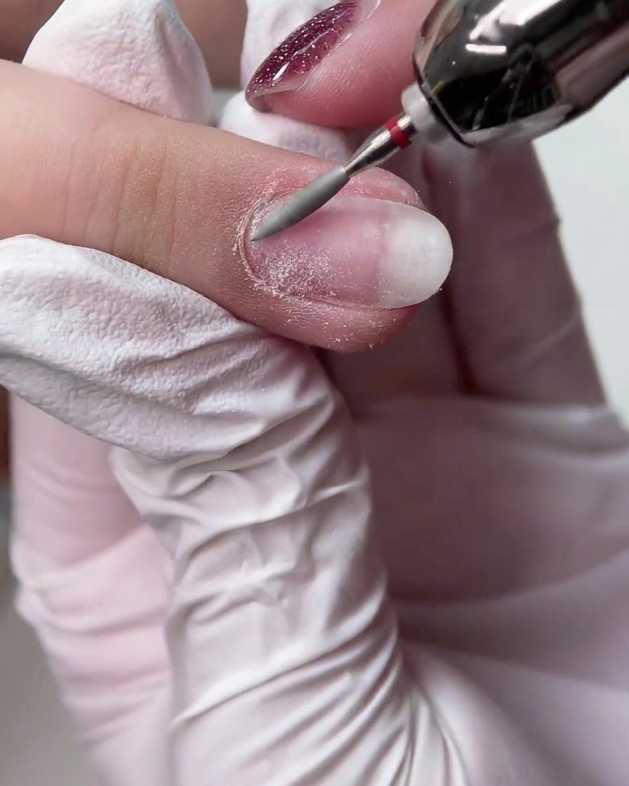 A gloved hand using an E-file with a long tapered bit to smooth the surface of a natural fingernail.