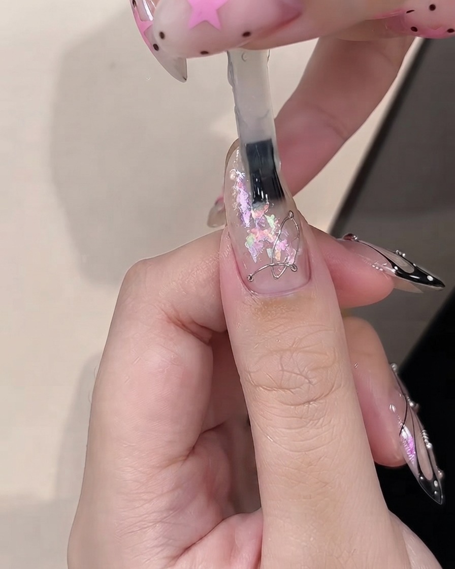 A brush applying builder gel over decorative opal pieces embedded on a clear-coated nail.