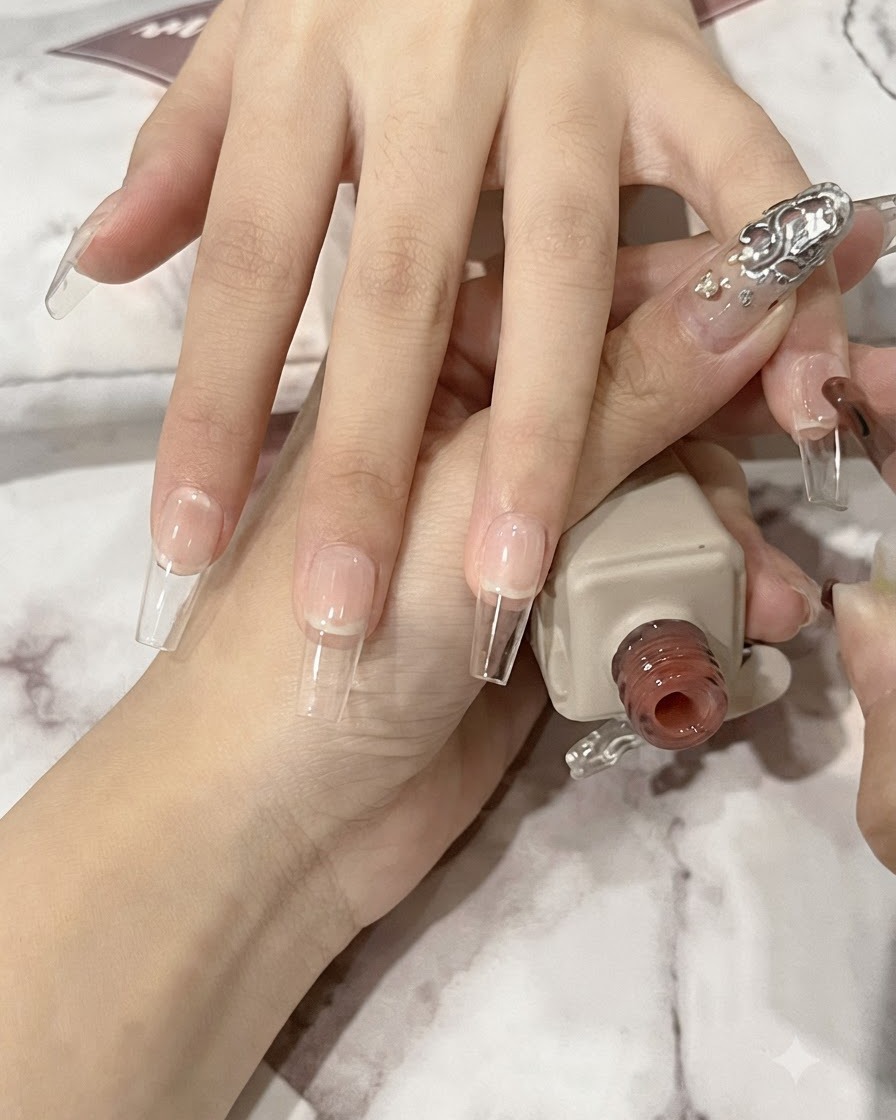 A nail brush applying a thin layer of translucent nude gel onto a long coffin-shaped natural nail plate.