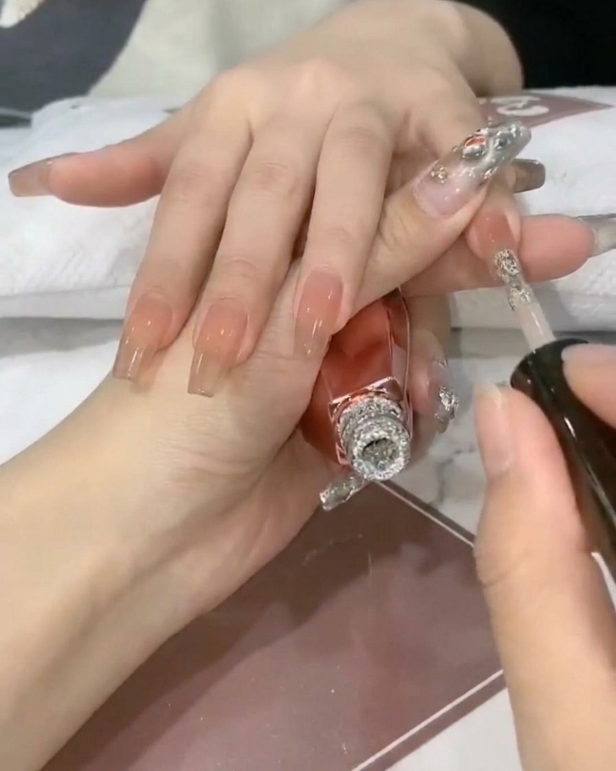 A nail brush depositing sparkling silver glitter gel onto the tip of a translucent nude nail.