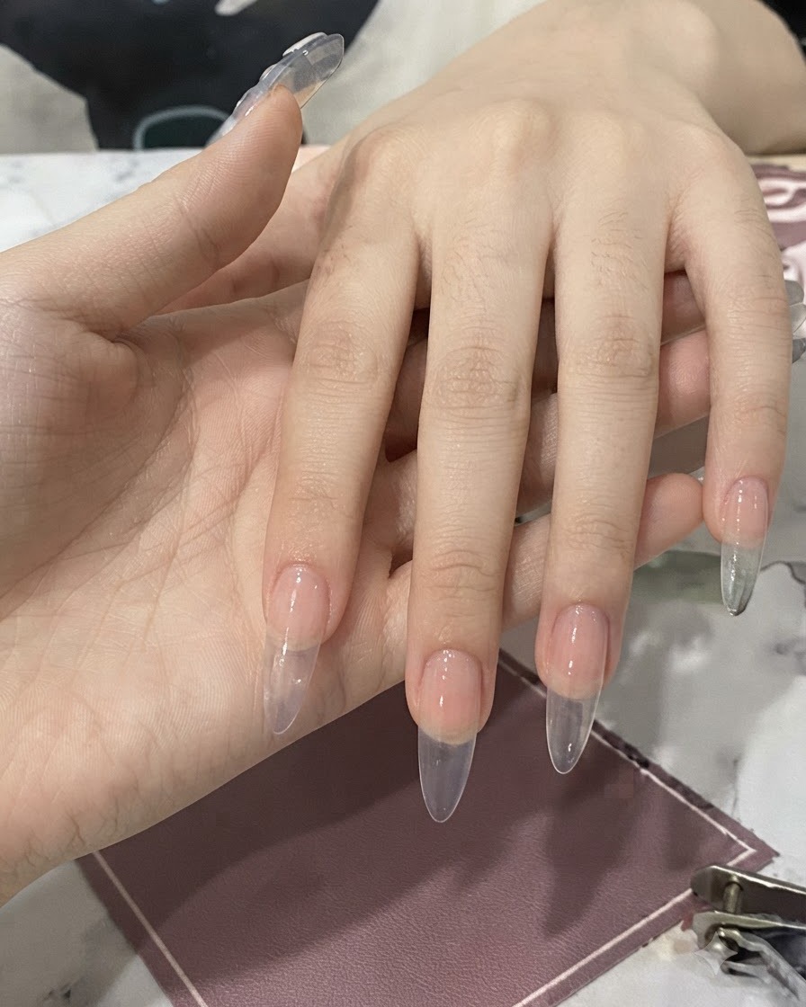 A hand holding a bottle with a brush applying clear nail glue onto a prepped fingernail.