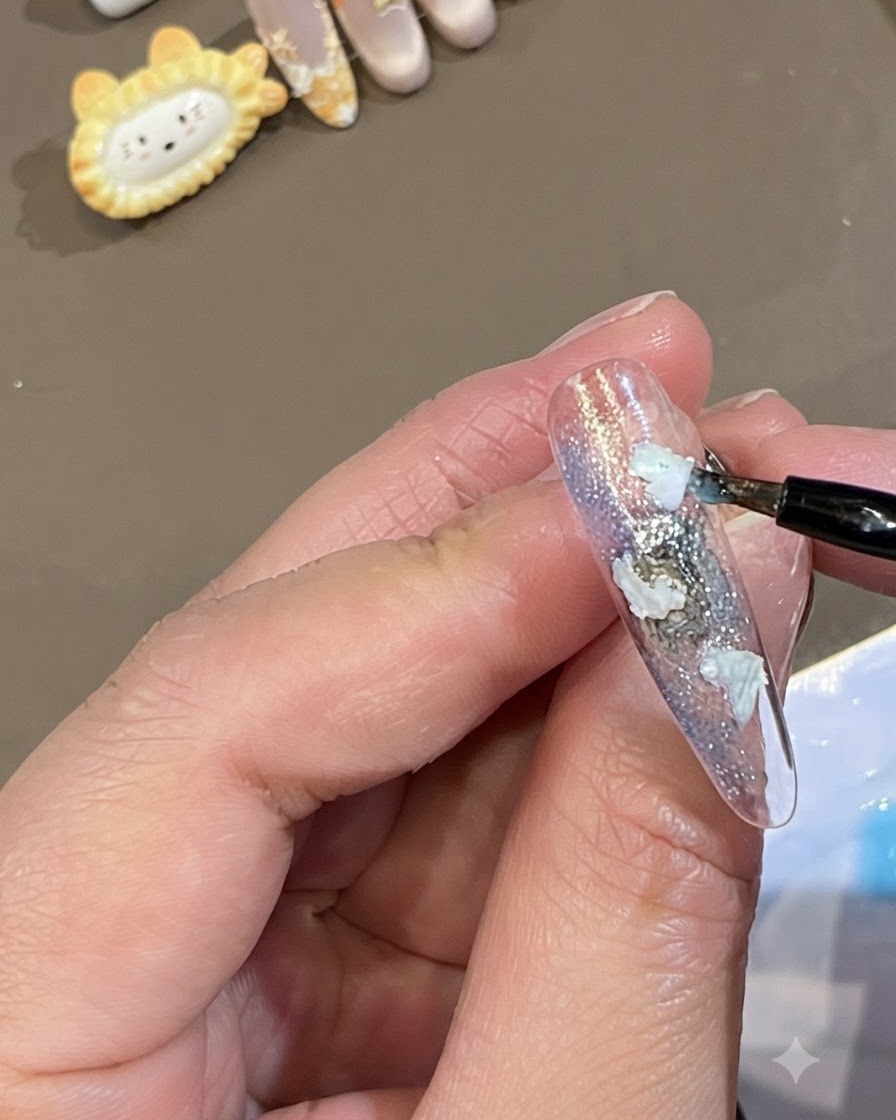 A fine brush dotting translucent blue gel around the edges of white shell pieces on a nail.