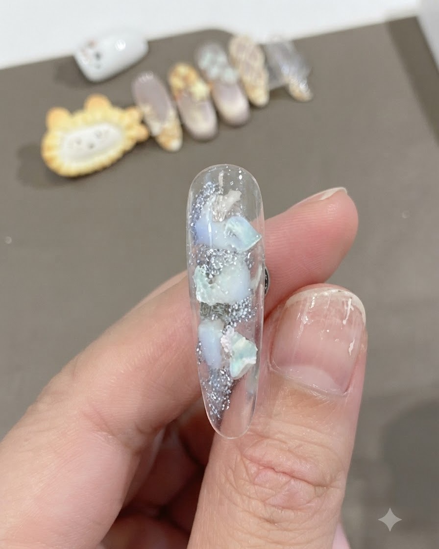 A close-up of a clear long nail featuring embedded shell pieces and shimmering texture with a soft white gradient blend.