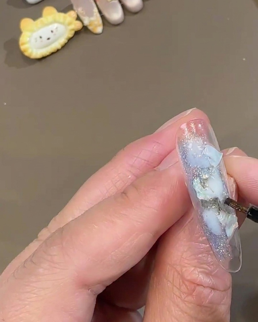 A fine detail nail brush dabbing white sparkling fairy powder onto a clear press-on nail decorated with blue shell pieces.