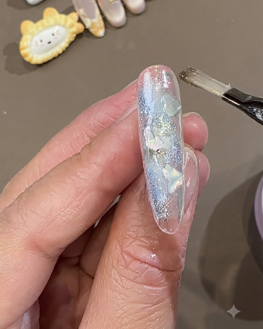 A flat gel brush applying a thick layer of clear soft encapsulation gel over a nail decorated with shell pieces and glitter.