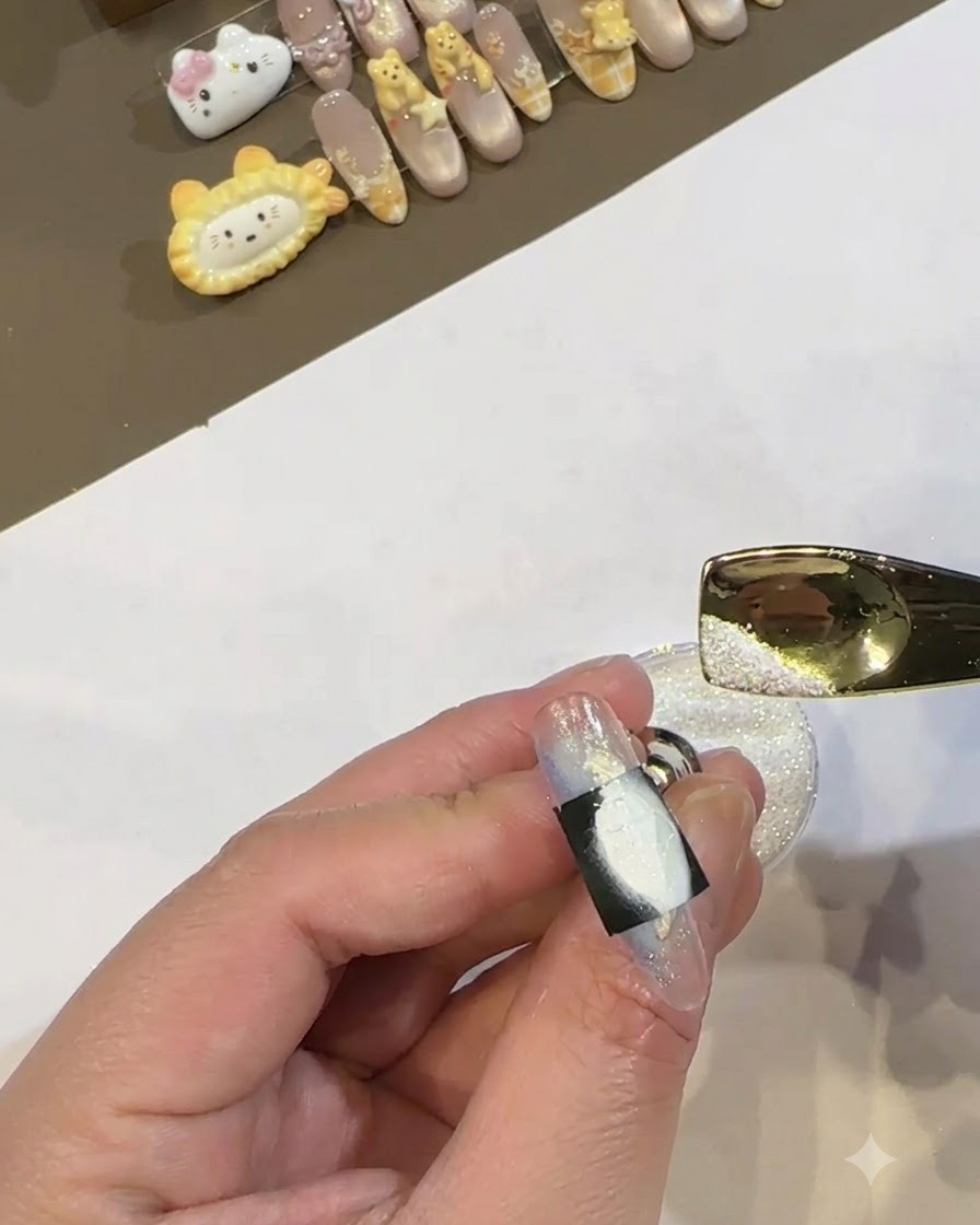 A gold metal scoop pouring shimmering white powder over the wet white gel area framed by a black stencil on a nail.