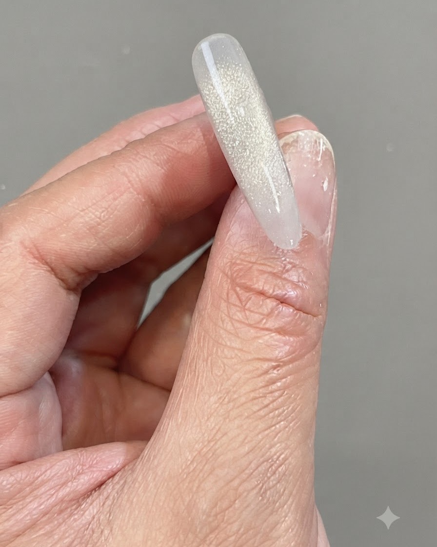 A hand holding a clear nail tip featuring a shimmering, vertical gradient made of reflective shattered diamond gel.