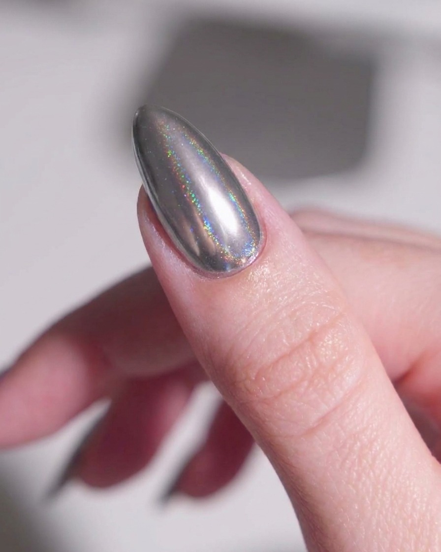 A soft makeup brush gently sweeping away loose metallic silver powder from the skin around an almond-shaped chrome nail.