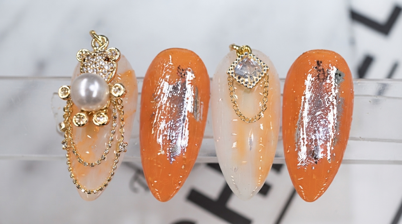 Close-up of a finished set of Japanese style press-on nails featuring an orange and white gradient base, silver transfer foil, and a 3D gold bear charm with a pearl and rhinestone chains.