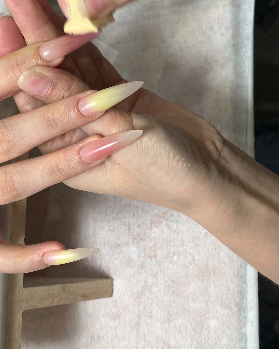 A hand being worked on with a sponge blending green and coral red tones onto long almond nails.