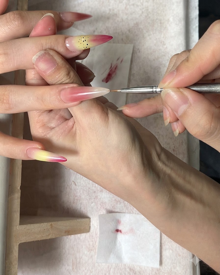 A fine nail art brush applying brown dots as stamens onto a gradient fingernail.