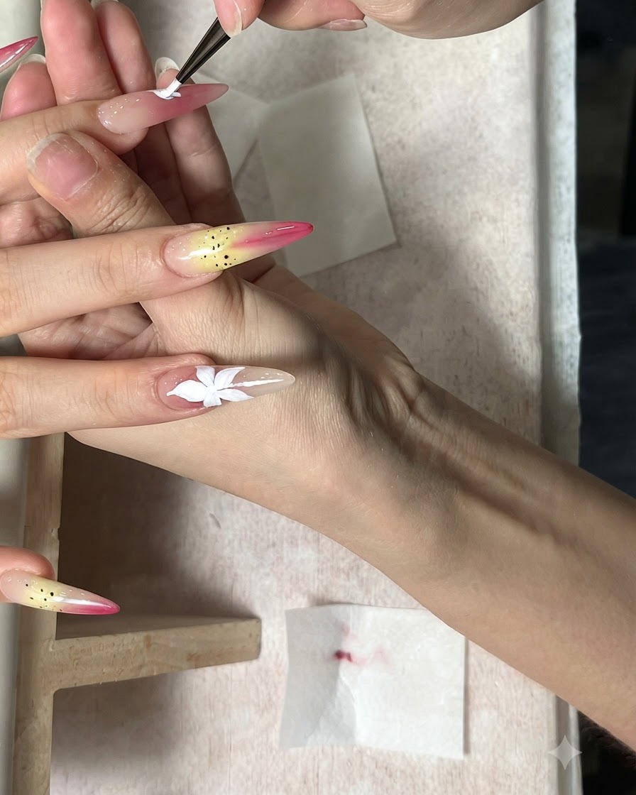 A fine brush painting a white five-petal flower on an elongated nude nail.
