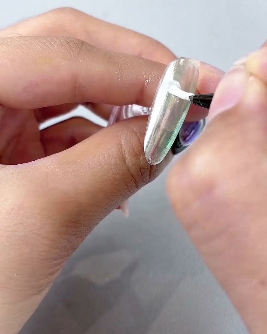 A fine nail art brush applying white painting gel in curved petals on a silvery chrome nail surface.