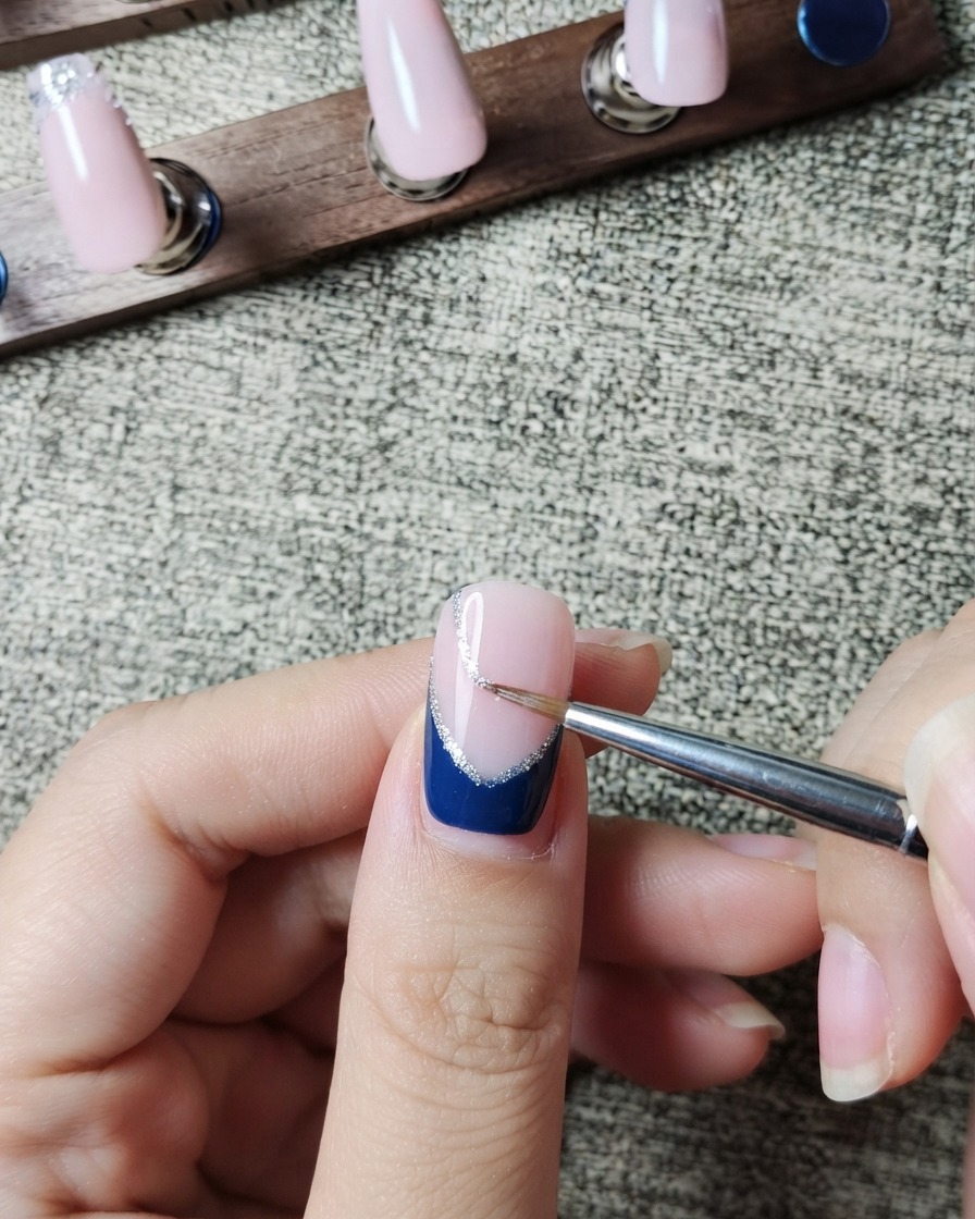 A liner brush carefully painting a thin silver glitter line along the inner edge of a dark blue French tip.