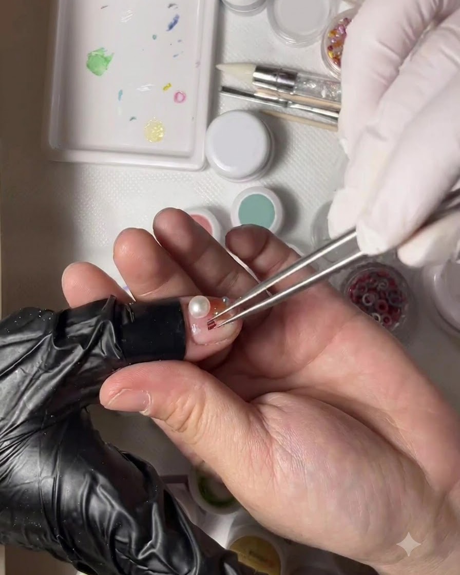 A gloved hand holding tweezers placing a tiny gold bead next to a pearl on a decorated thumb nail.
