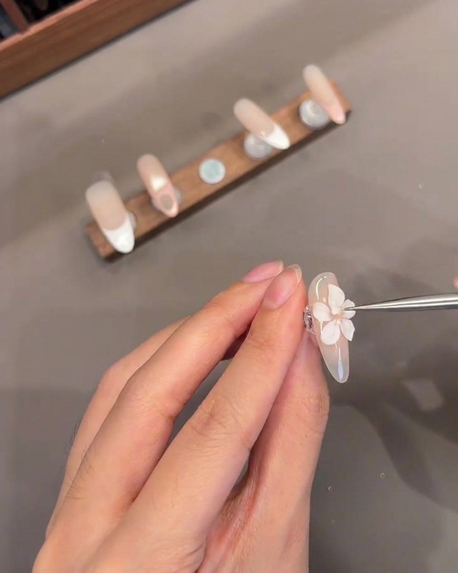 A close-up of a thin tool adding a small white gel center to a sculpted 3D flower on a fingernail.