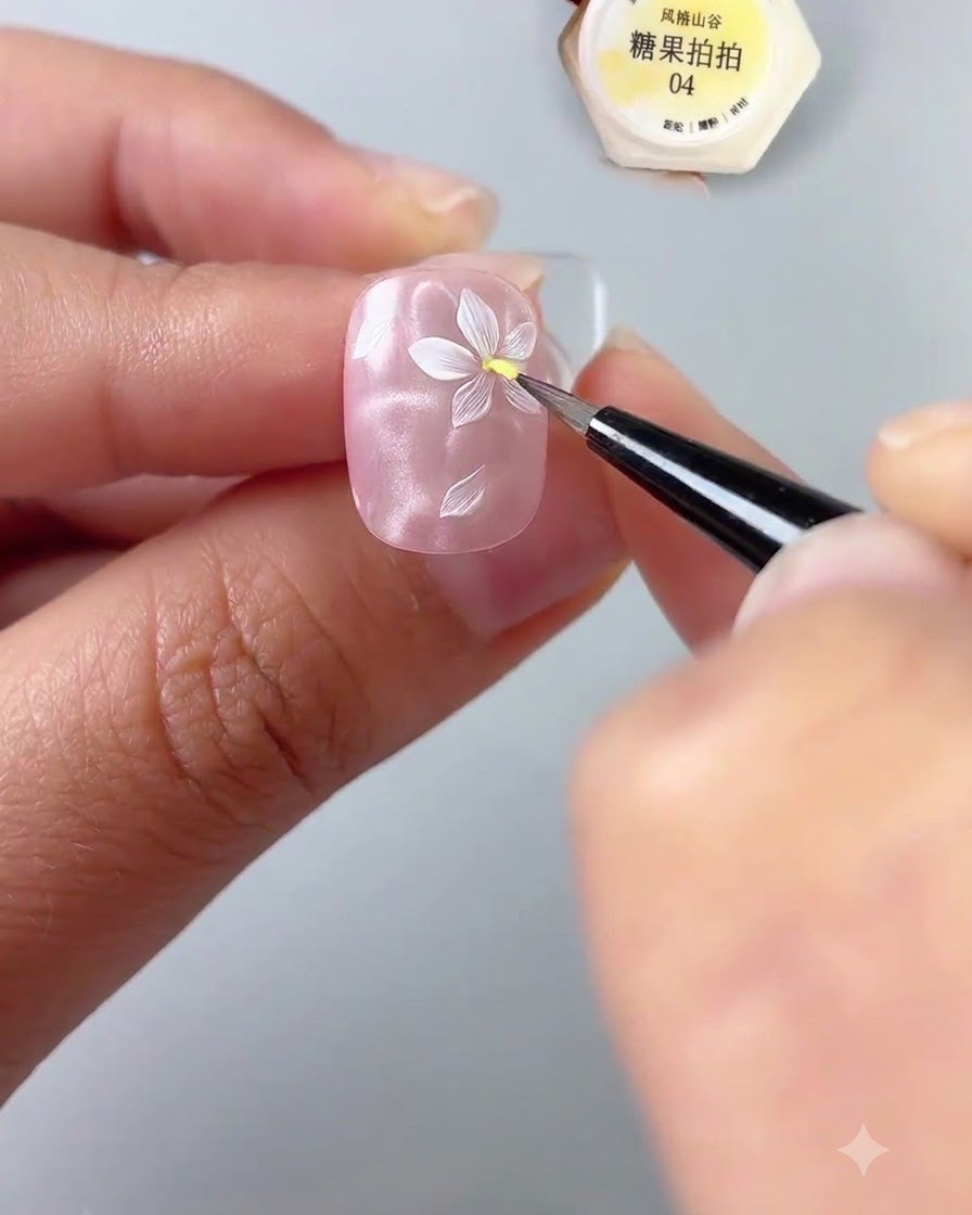 A close-up of a hand holding a nail while using a fine detail brush to apply a dot of yellow gel to the center of a painted white flower on a glossy pink nail base.