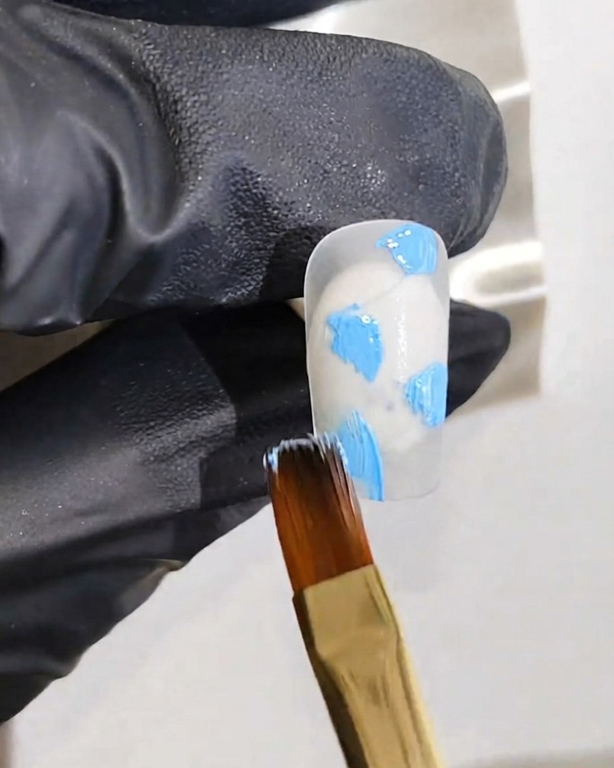 A gloved hand holding a fine art brush applying blue gel dots onto a white nail base.