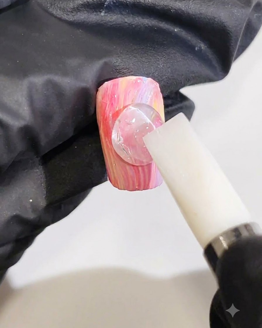 A gloved hand using a white silicone tool to shape a clear drop of 3D gel into a ripple line on a pink and yellow marble nail.