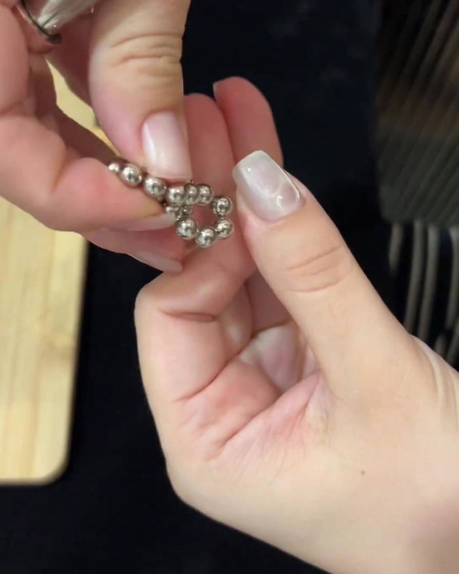 Hands using magnetic tools on both sides to manipulate metallic beads and create water ripples on the nail.